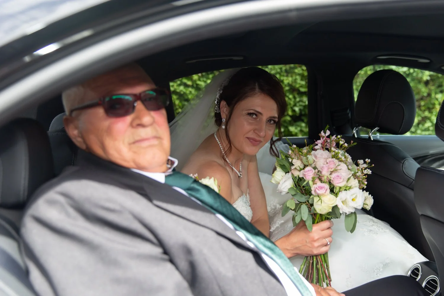A bride holding a bouquet of pink and white roses sitting in a car with an older man, both dressed in wedding attire, with greenery outside the car window.