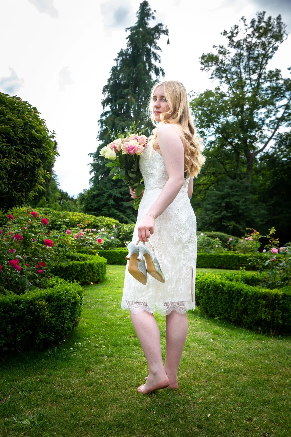 Woman in a white lace dress holding a bouquet of pink and white roses, carrying silver high-heeled shoes, standing barefoot on grass in a garden with rose bushes and tall trees.