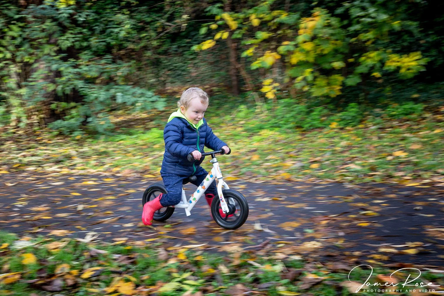 Young boy riding a balance bike through a wooded area with fallen leaves on the path in autumn.