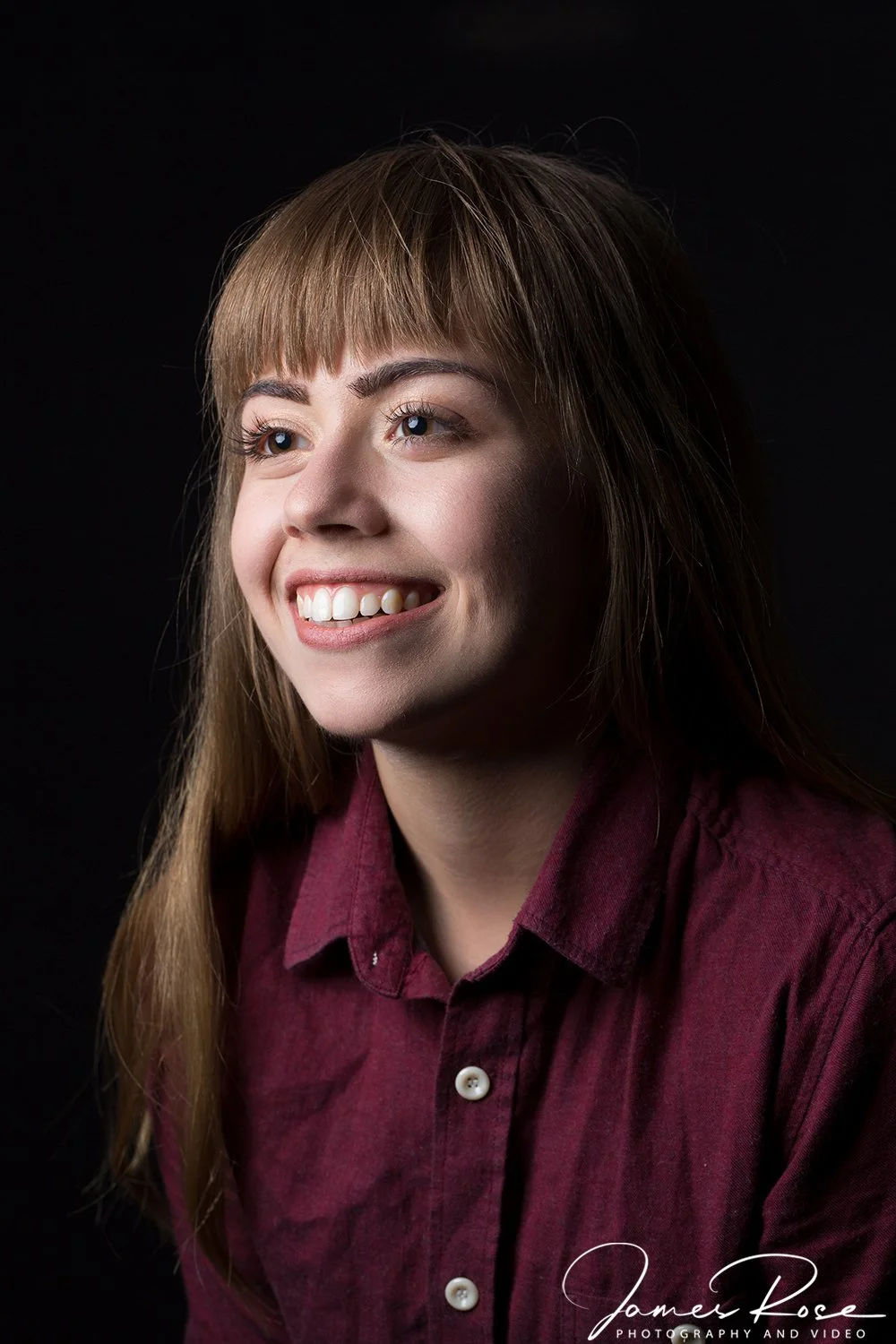 Portrait of a young woman with long brown hair, smiling, wearing a maroon shirt, against a dark background.