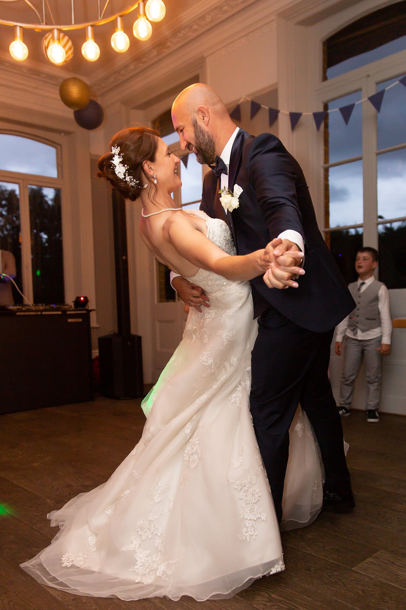 A bride and groom dancing closely during their wedding reception indoors, with a young boy in the background watching.