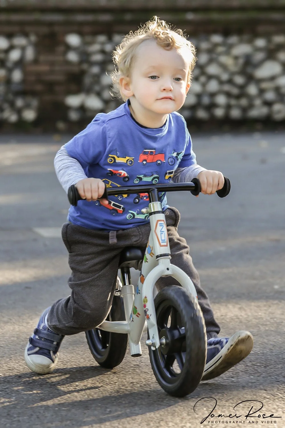 A young boy riding a balance bike outdoors on pavement, wearing a blue shirt with colorful vehicle graphics and dark grey pants, with a stone wall in the background.