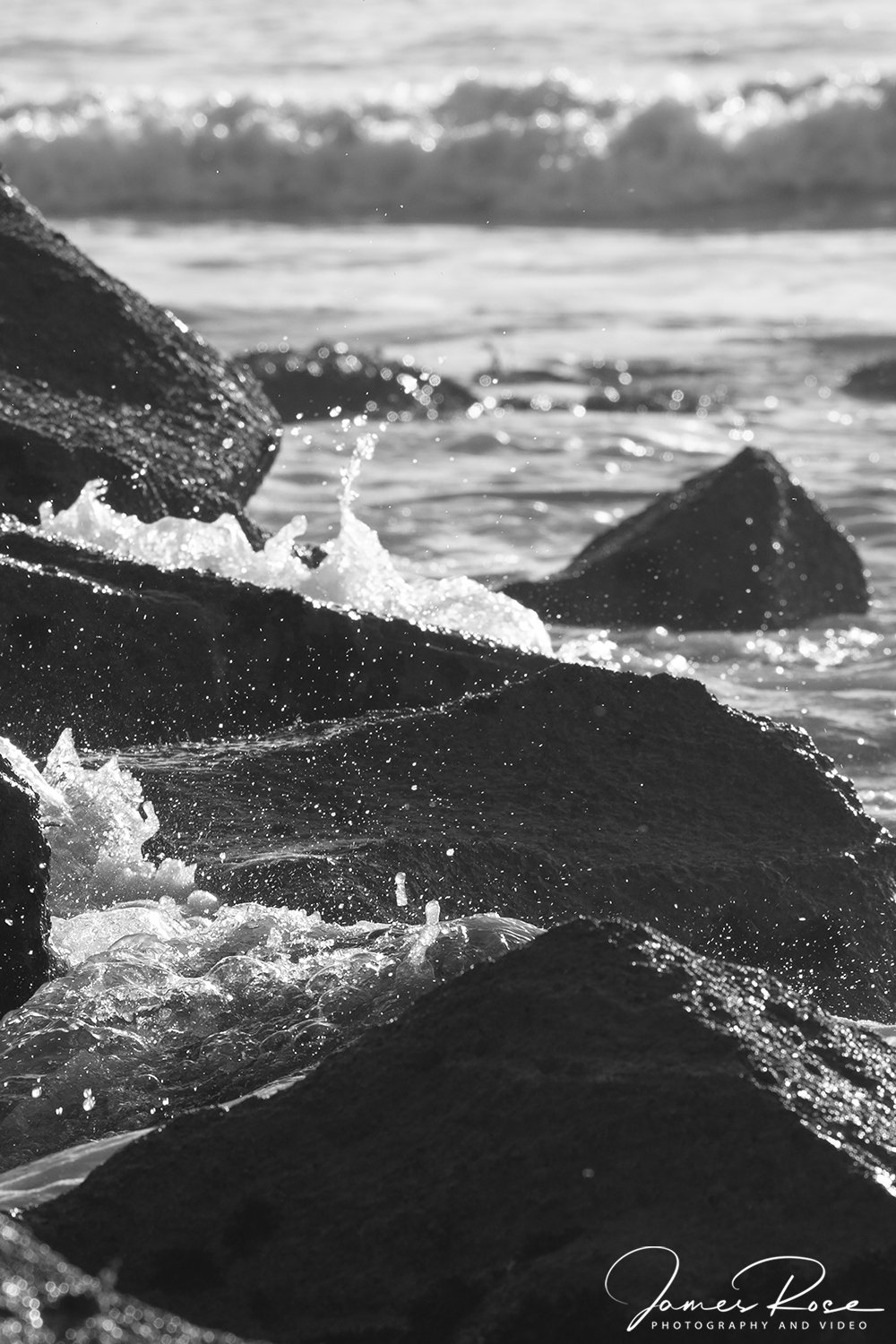 Black and white photo of ocean waves crashing against rocks near the shoreline.