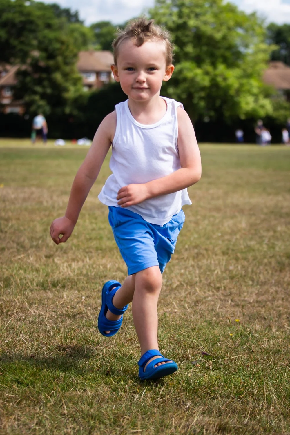 A young boy running on a grassy field outdoors, wearing a white sleeveless shirt, blue shorts, and blue sandals, with green trees and houses in the background.