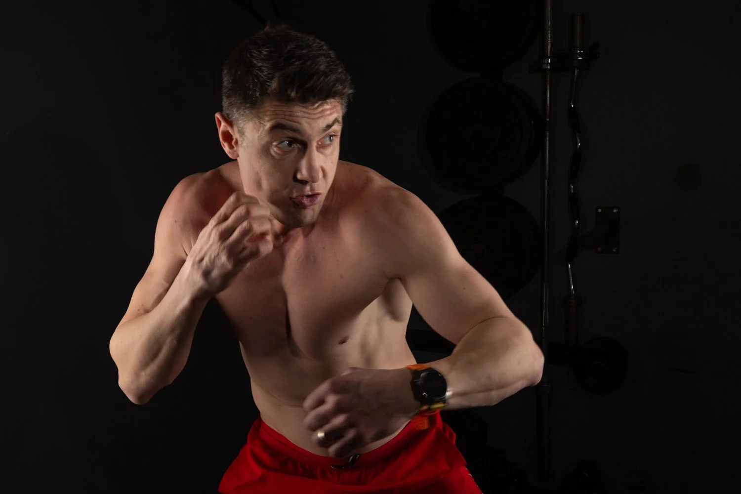 A shirtless man in red shorts prepares to punch, standing in a gym with weights in the background.