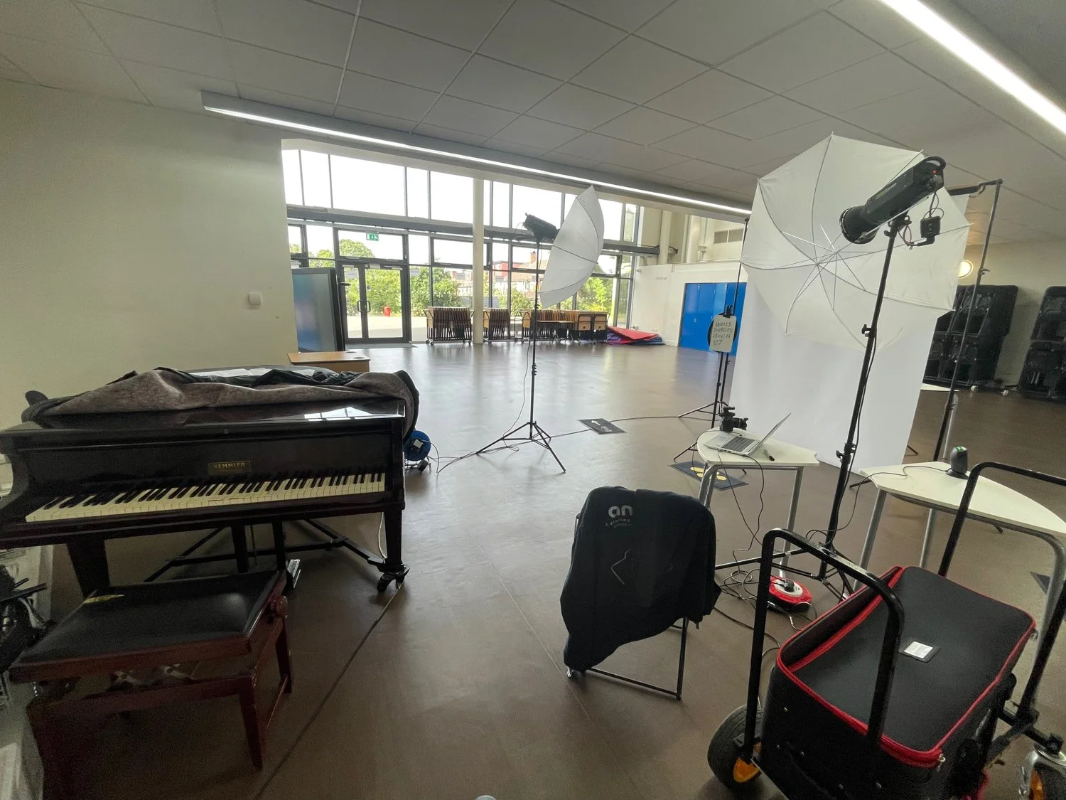 Indoor photography setup with a grand piano, tripods with studio lights, large umbrellas, a laptop on a small white table, and various photography equipment in a spacious room with large front windows and chairs outside.