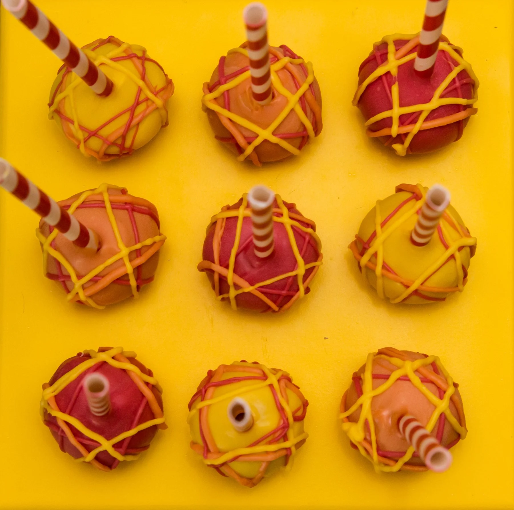 Colorful cake pops with yellow, pink, and orange icing, each with a striped straw inserted, arranged on a yellow surface.