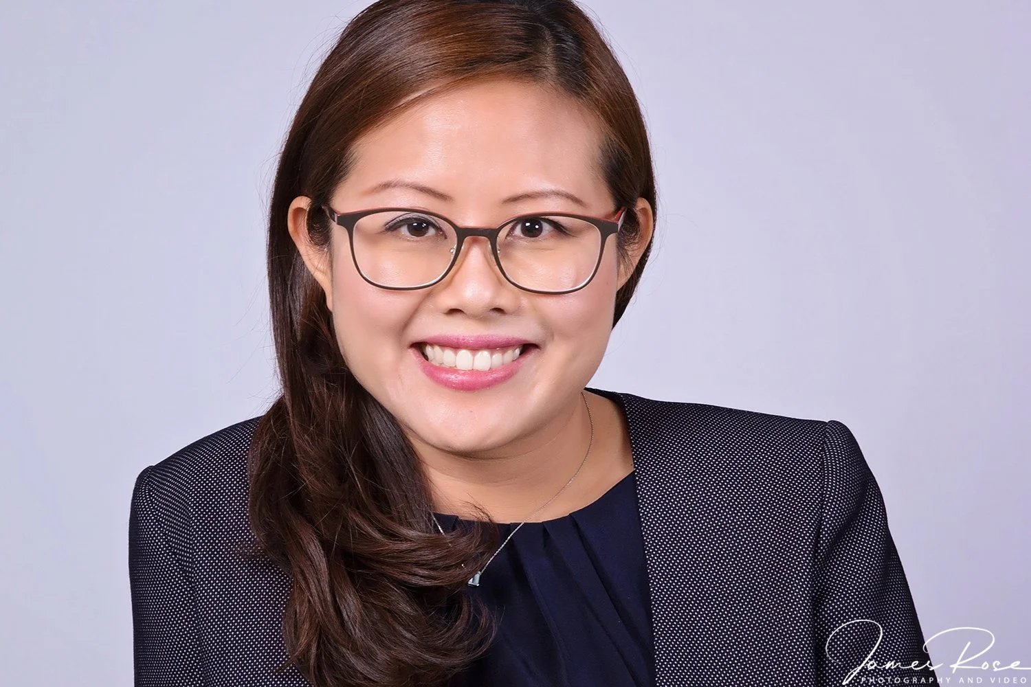 Portrait of a smiling woman with glasses, brown hair, wearing a dark blazer, against a light background.
