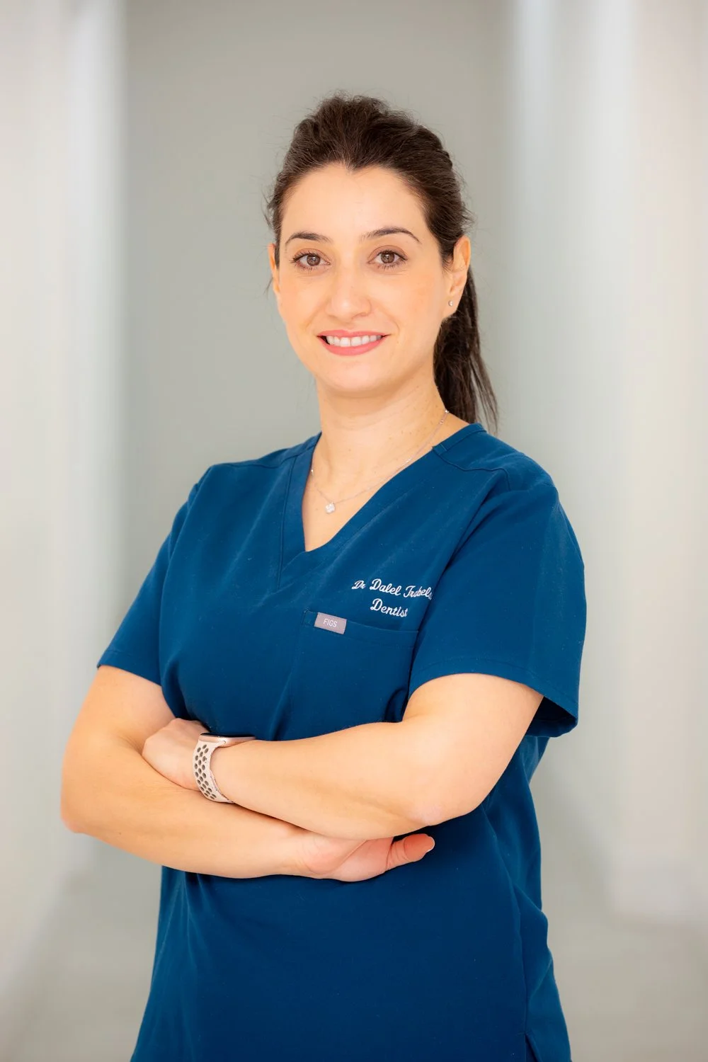 A woman in blue dental scrubs standing with arms crossed, smiling, in a professional setting.