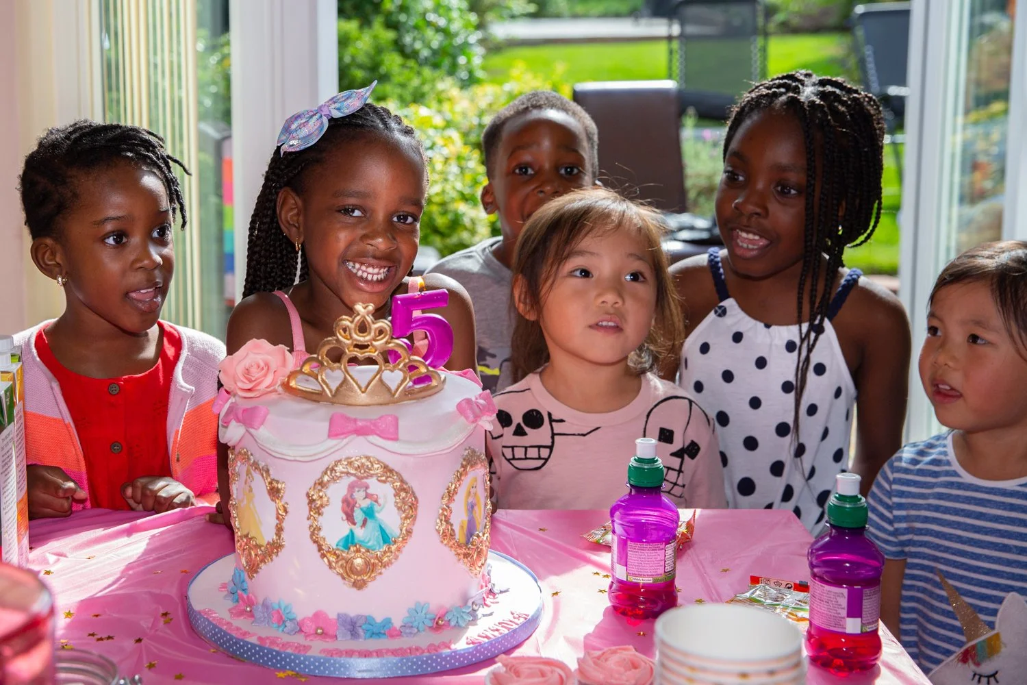 A group of children celebrating a birthday around a decorated cake with a princess theme, pink fondant, and a number 5 candle.