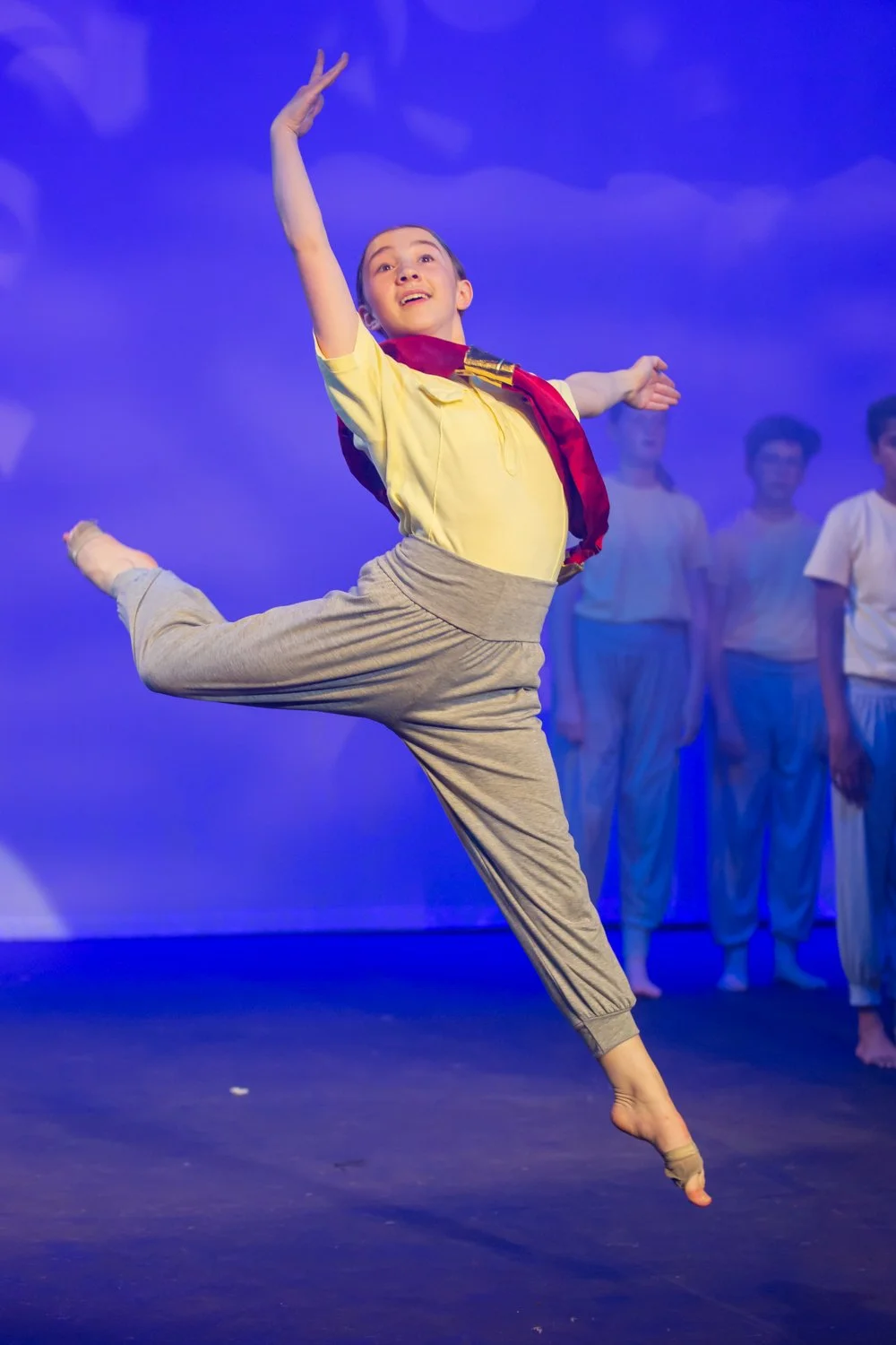 Young boy performing a dance move on stage with a bright blue background, wearing a yellow shirt, tan pants, and a red scarf, with other boys in white shirts in the background.