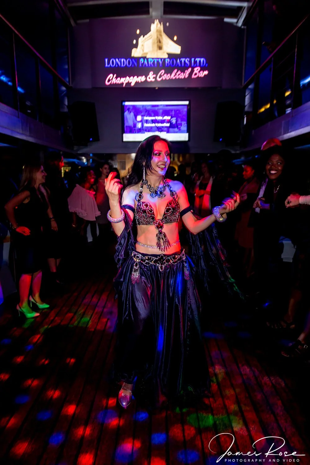 A woman dancing at a nightclub, wearing a black and red belly dance costume with jewelry and a dark background with guests and a LED screen displaying 'London Party Boats Ltd. Champagne & Cocktail Bar'.