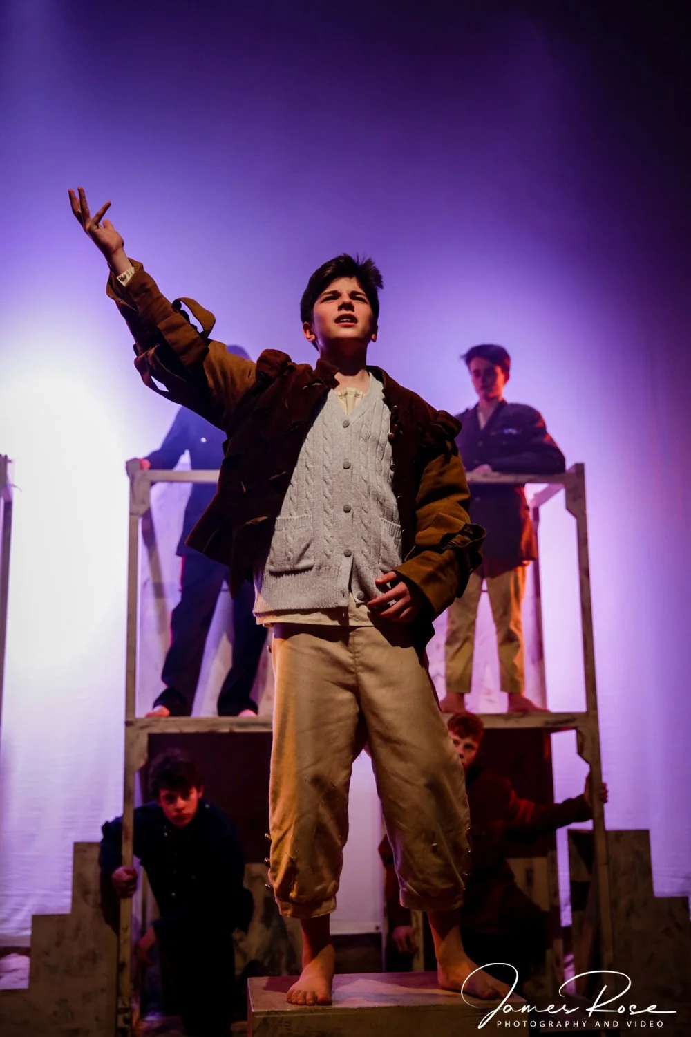 Young boy performing on stage with other children in the background, some on a wooden structure, under purple stage lighting.