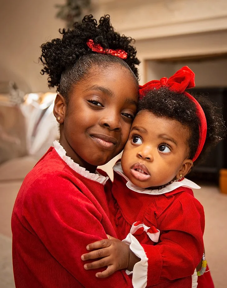 A young girl and a toddler girl wearing matching red outfits embrace each other indoors.