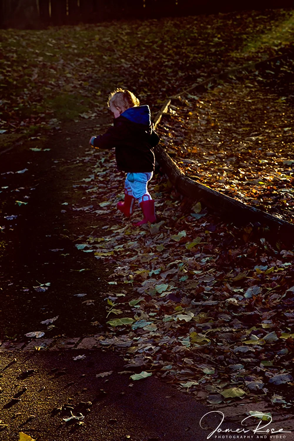 A young child wearing red rain boots, a black jacket, and blue pants with red accents, walking along a leaf-covered dirt trail bordered by a wooden log, during sunset in an outdoor setting.