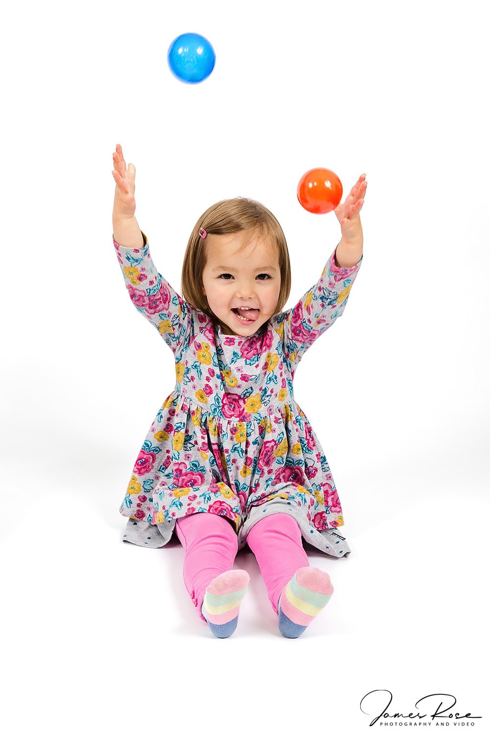 A young girl sitting on the floor with her legs extended, wearing pink and rainbow socks, a floral dress, and playing with two colorful balls, one blue and one orange.