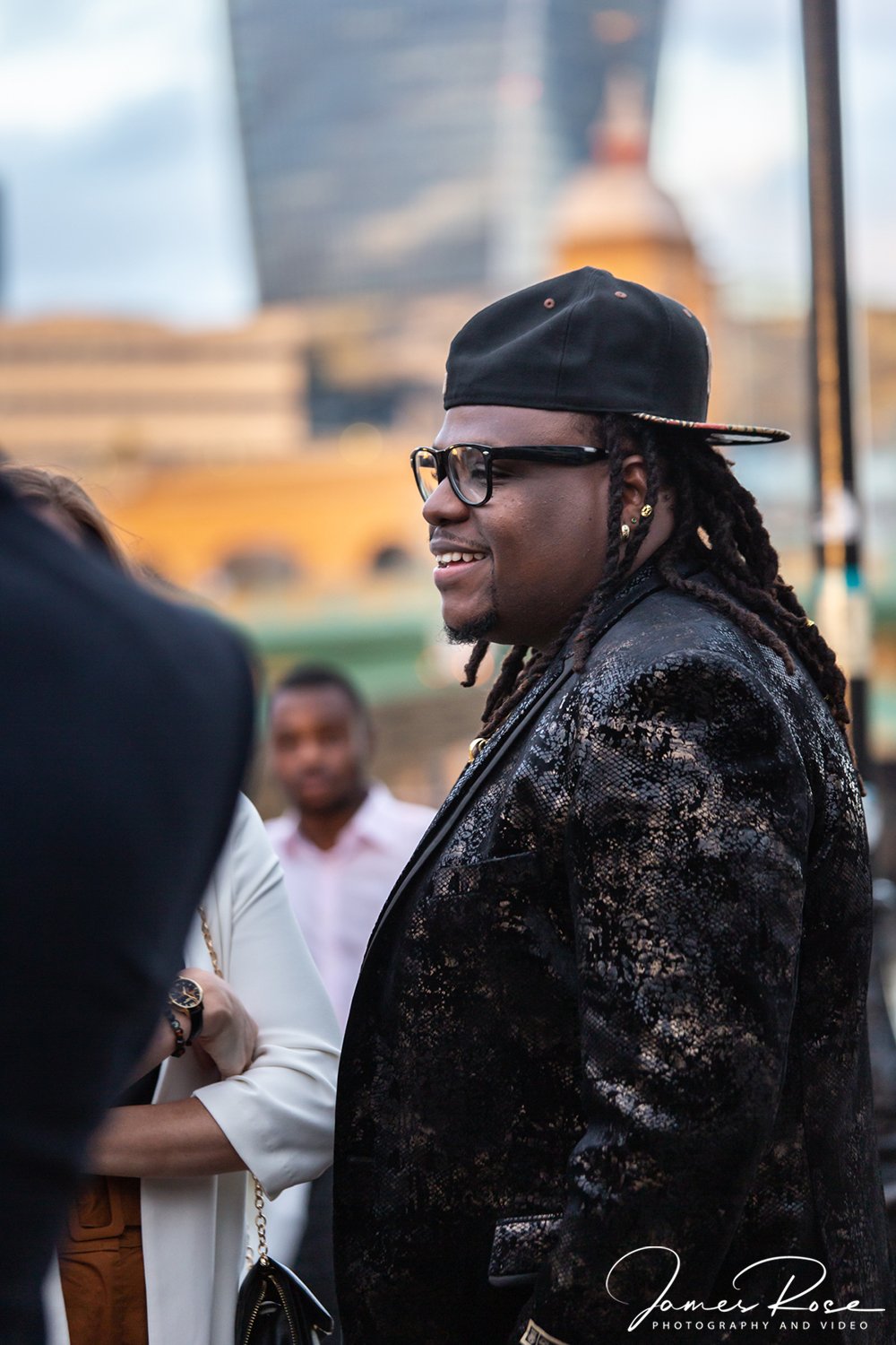 A man with dreadlocks wearing a black baseball cap backwards, black glasses, and a black and gold patterned blazer, smiling during an outdoor event.