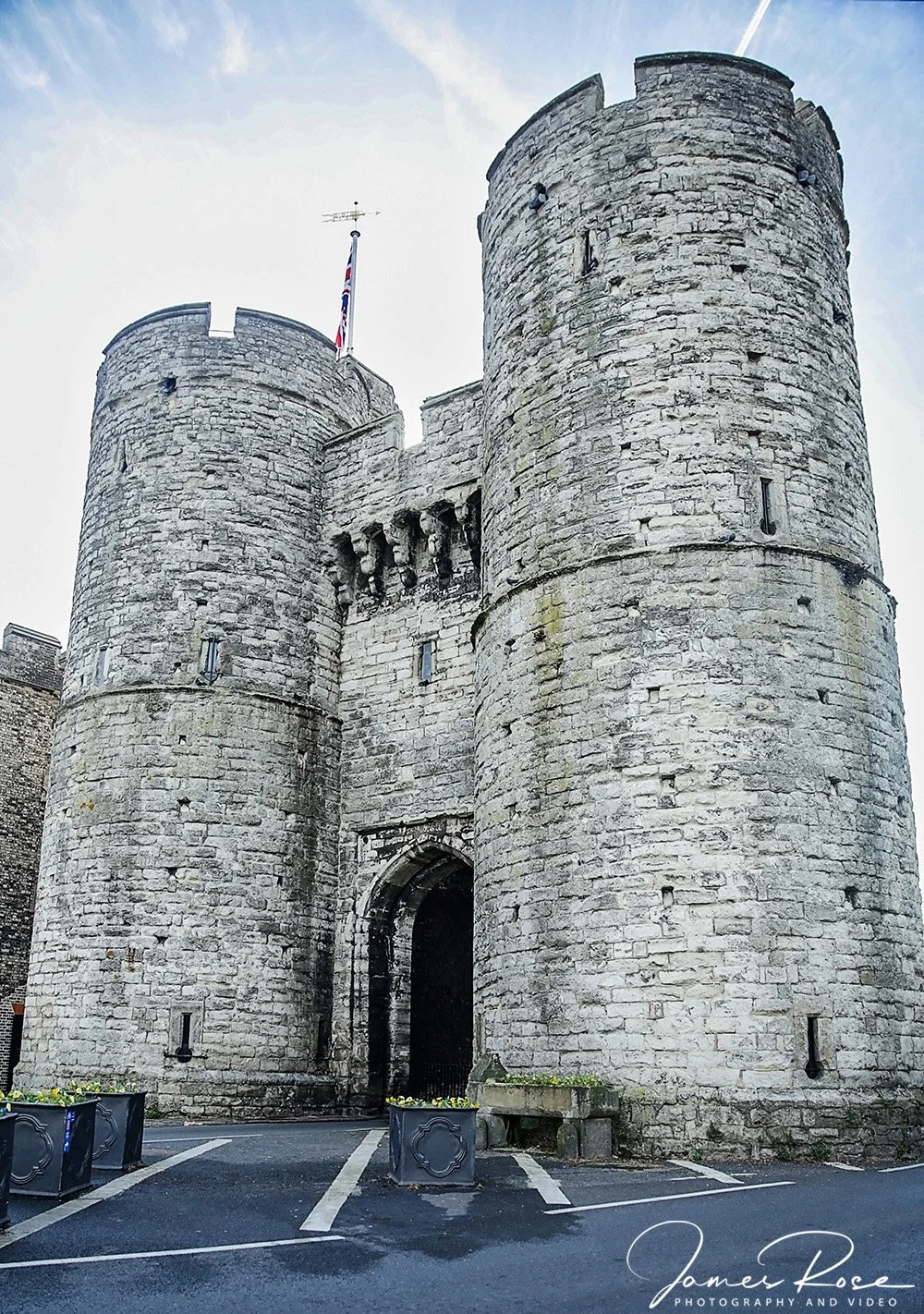 A historic stone castle with two large round towers on each side, a narrow entrance arch, and a flag on top of the left tower. The sky is cloudy with a faint rainbow visible, and there are planters in front of the castle.