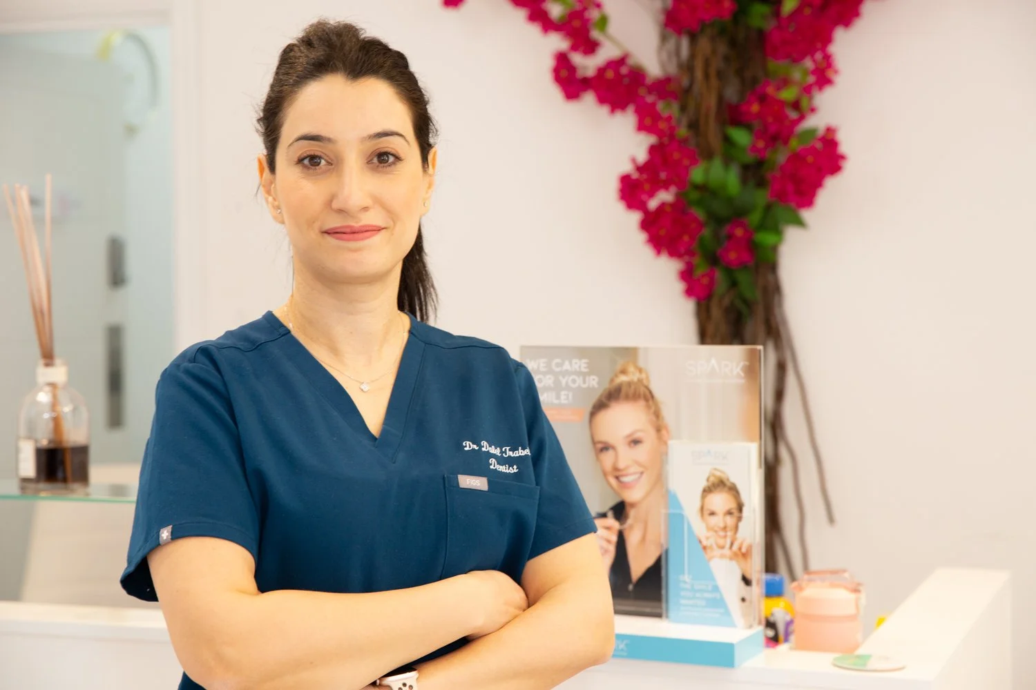 A woman wearing navy medical scrubs with a name badge, standing with arms crossed inside a dental clinic. Behind her, there is a pink flowering plant, a brochure stand with promotional materials, and a clear container with reed diffusers.