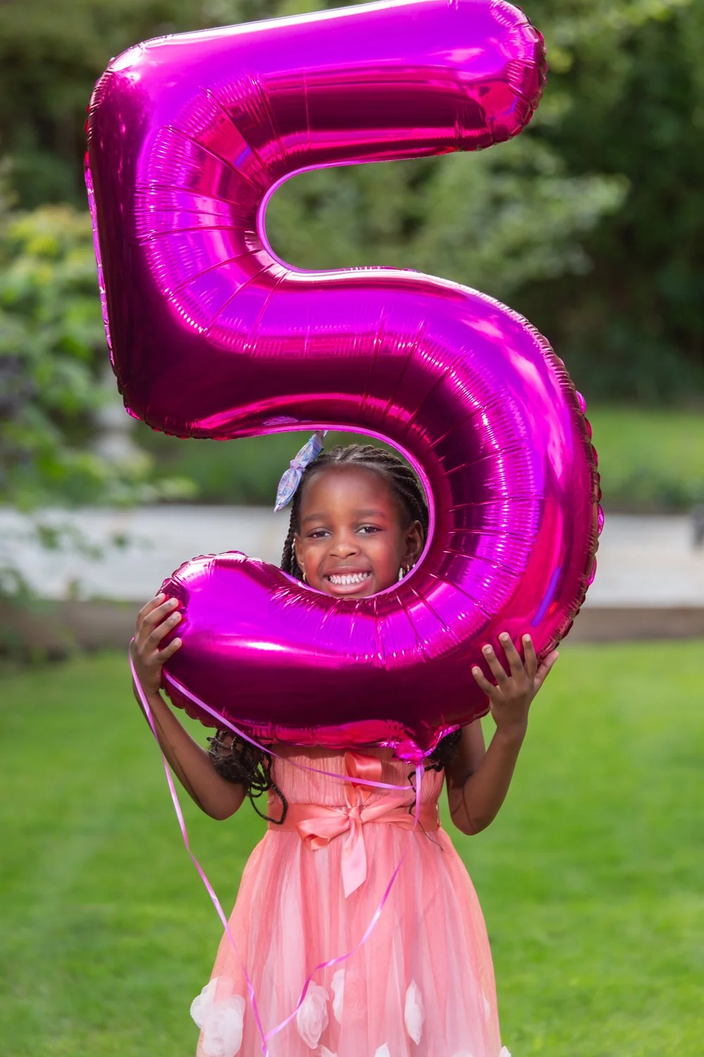 A young girl with braids and a light pink dress is holding a large pink foil balloon shaped like the number 5, smiling at the camera outdoors with greenery in the background.