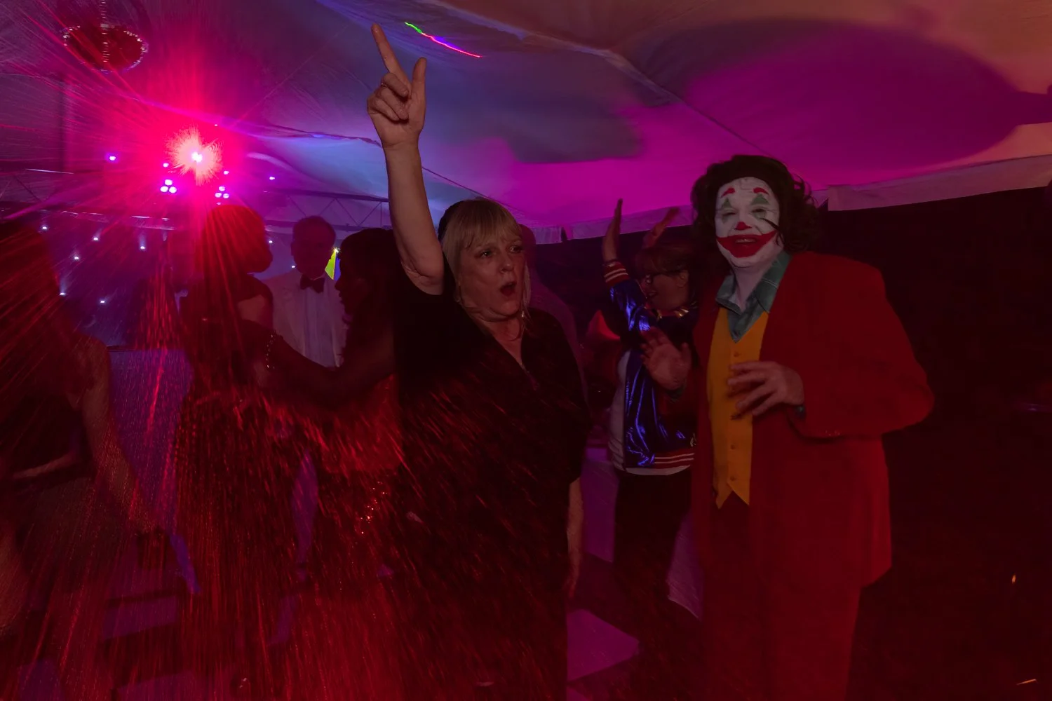 People dancing and celebrating at a party with colorful lights, including a woman with blonde hair raising her hand and a man dressed as a clown with white face paint, red hair, and a red jacket.