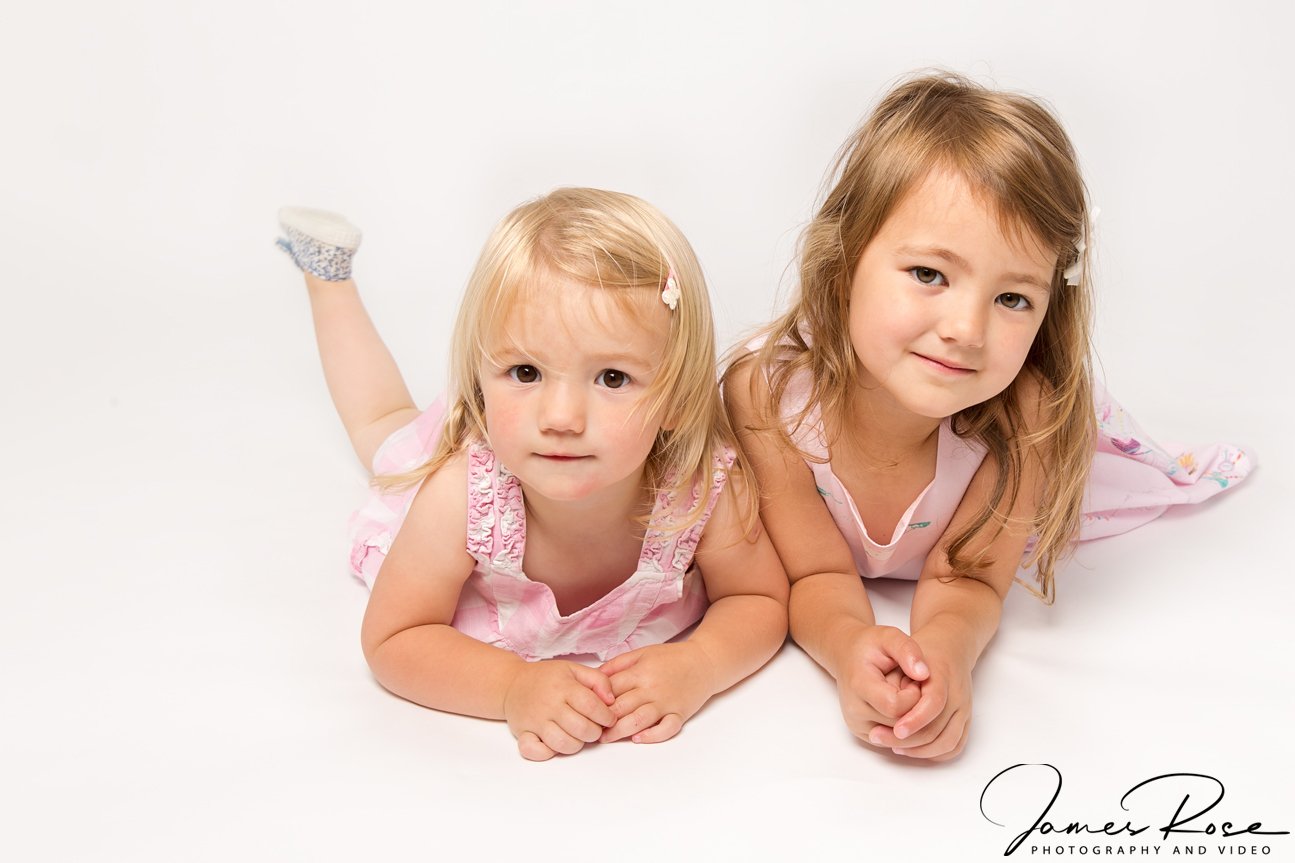 Two young girls with blonde hair lying on a white background, wearing pink dresses and looking at the camera.