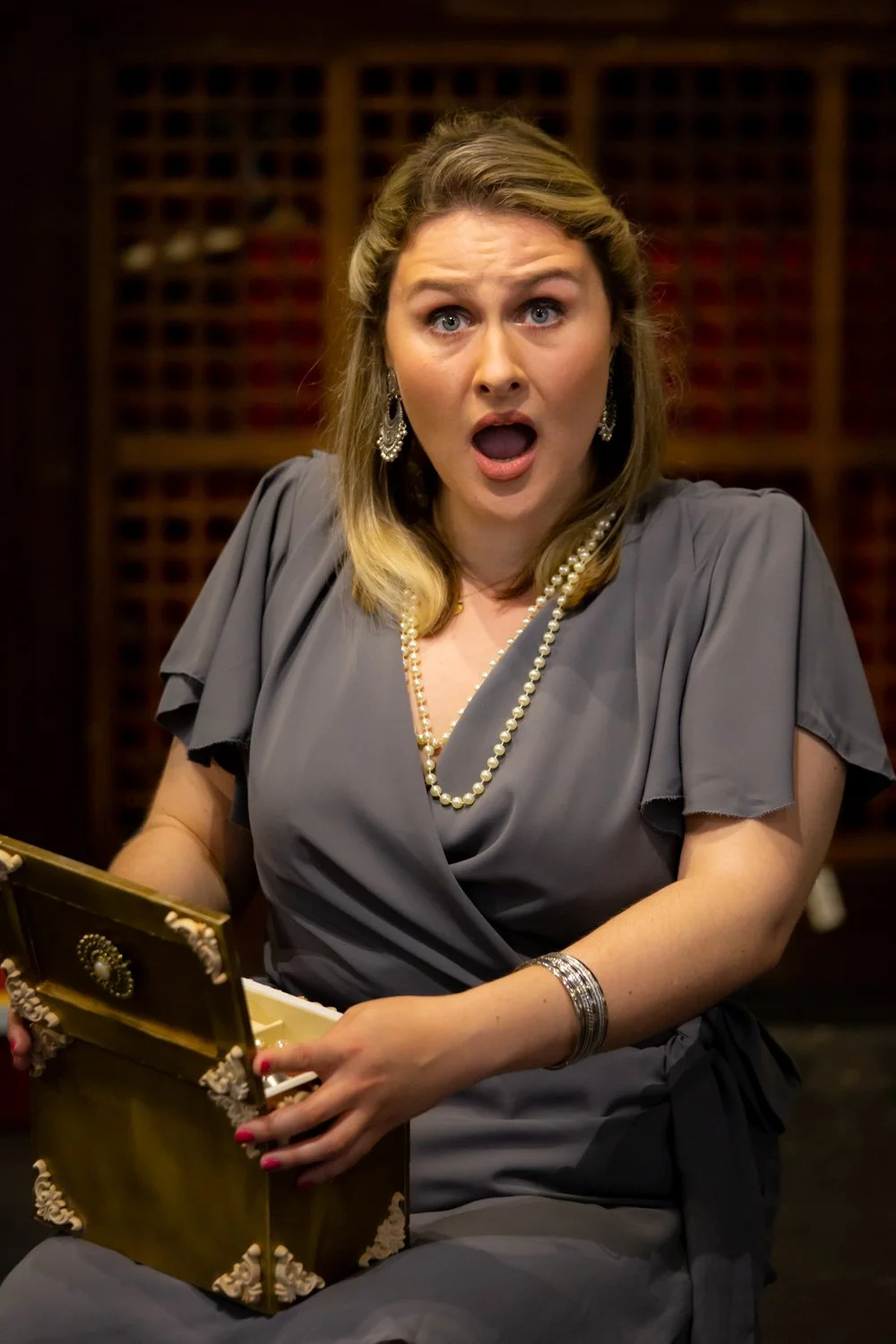 A woman with surprised expression, wearing a gray dress, pearl necklace, and earrings, holding an ornate box in a room with wooden shelves in the background.