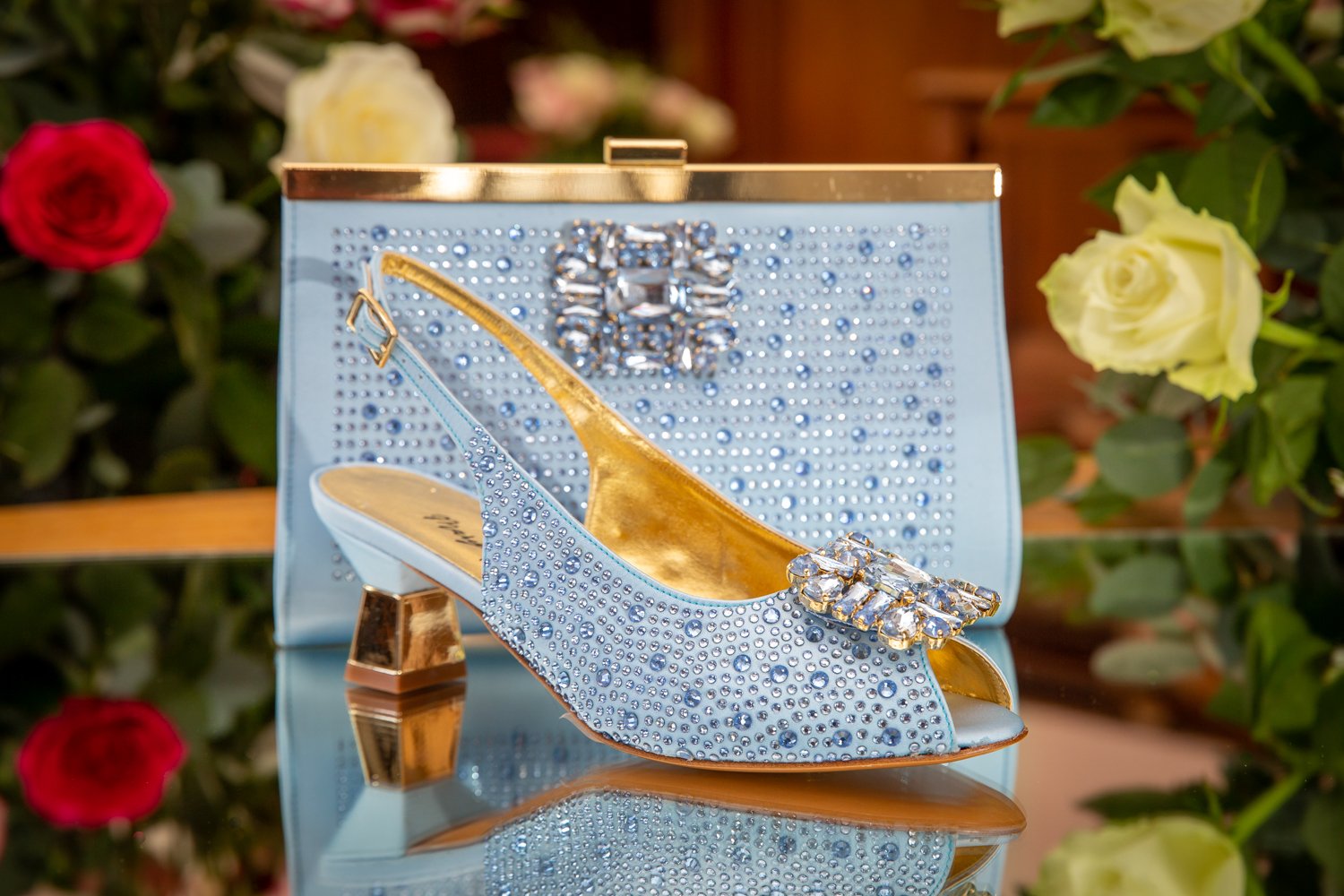 A blue high-heeled shoe adorned with rhinestones, placed in front of a matching decorative clutch purse with rhinestones, surrounded by colorful roses.