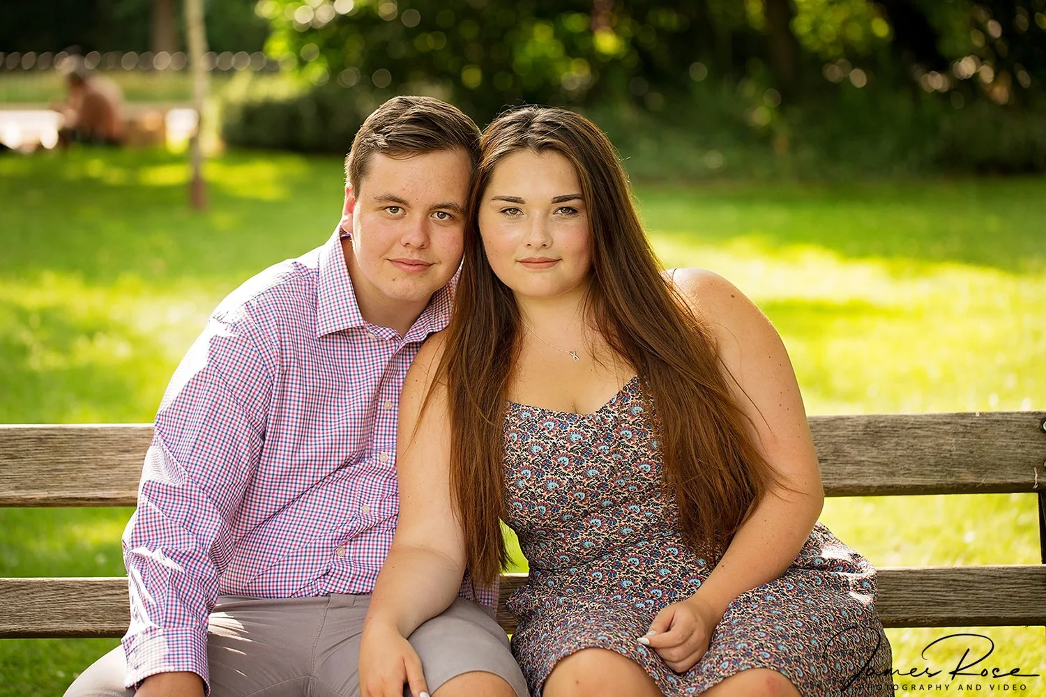 A young man and woman sitting on a wooden park bench outdoors, smiling at the camera in a grassy area with trees in the background.