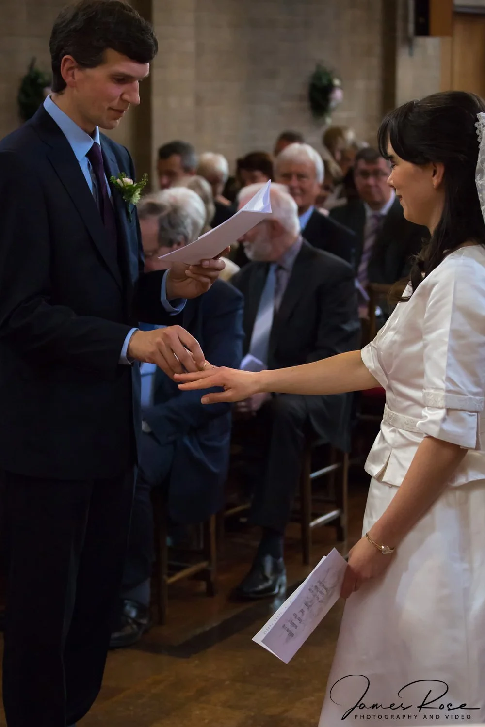 A man in a dark suit and a woman in a white wedding dress exchanging rings during a wedding ceremony with guests seated in the background.