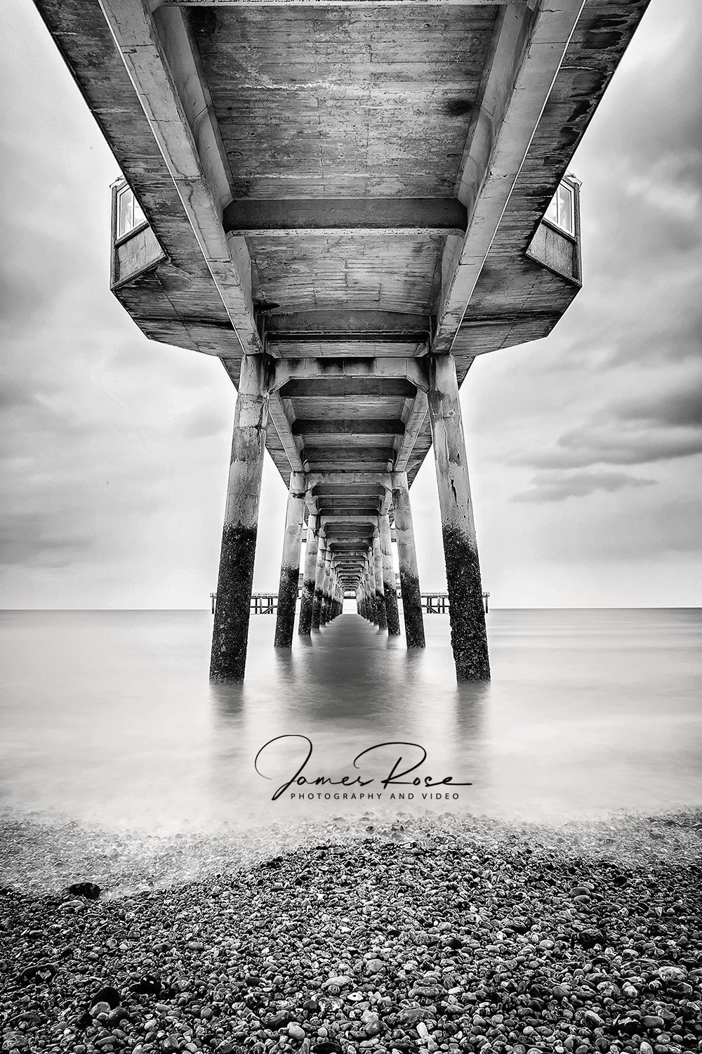 Underneath a pier extending over the ocean seen in black and white, with stone-covered beach in the foreground.