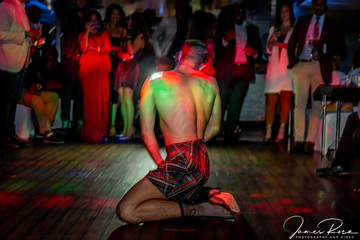 A man kneeling shirtless on the floor with his back to the camera, wearing a plaid kilt and fishnet socks, surrounded by a group of people dressed formally and casually at a party or celebration.