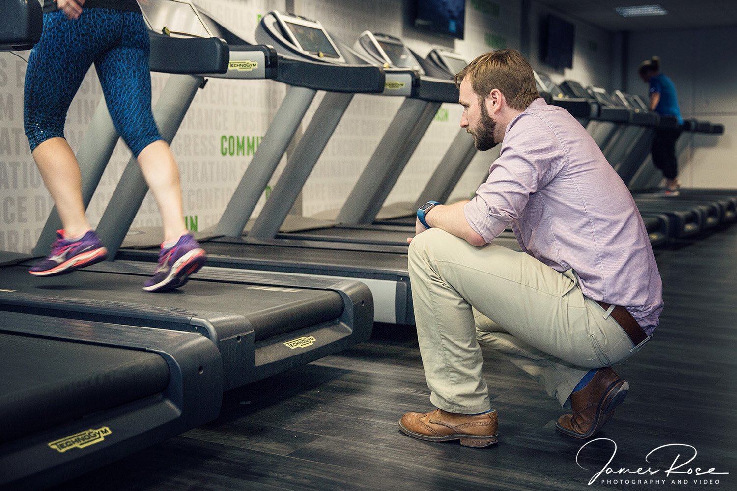 A man crouching and watching a woman run on a treadmill in a gym, with several other treadmills lined up in the background.