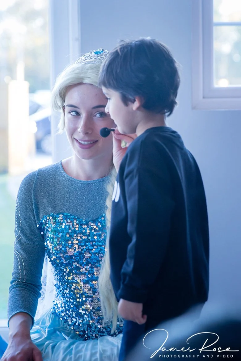 A young woman dressed as a princess, wearing a tiara and a sparkly blue dress, is smiling at a young boy who is holding a microphone close to her face.
