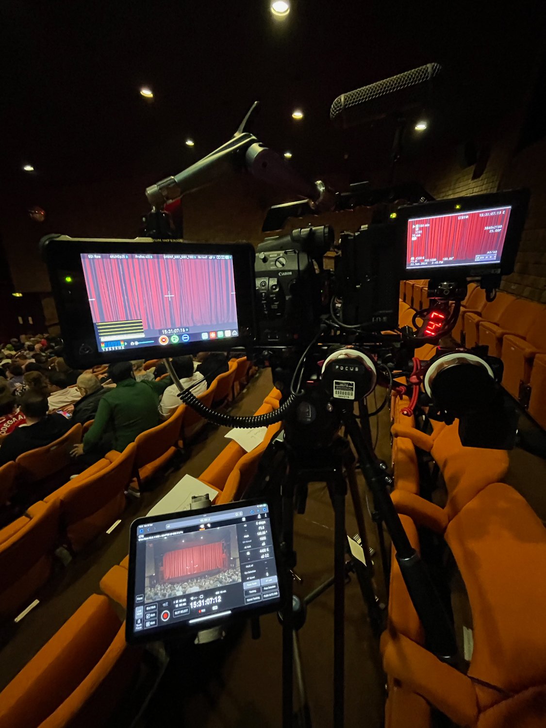 A camera setup capturing a scene at a theater or auditorium, with an audience seated in orange chairs facing a red curtain on stage.