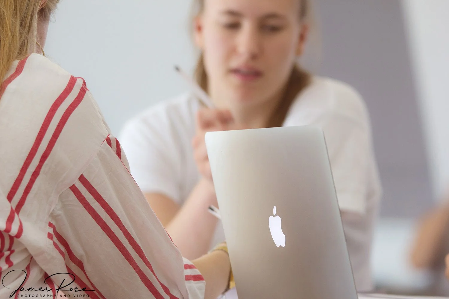 Two women are sitting at a table with a MacBook laptop, one facing away and the other mid-conversation, in a bright indoor setting.