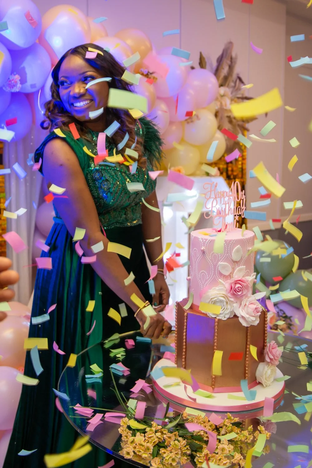 Woman celebrating her birthday with a tiered pink and gold cake decorated with flowers, surrounded by balloons and colorful confetti.