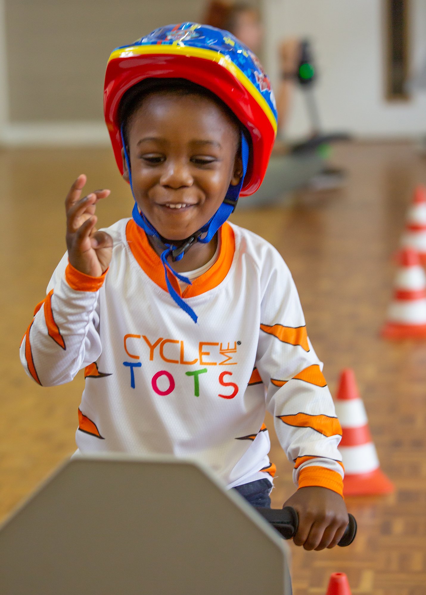 A young child wearing a colourful helmet and a white jersey with orange, green, red, and blue letters spelling 'CYCLE ME TOTS' is riding a scooter in an indoor space decorated with orange traffic cones.