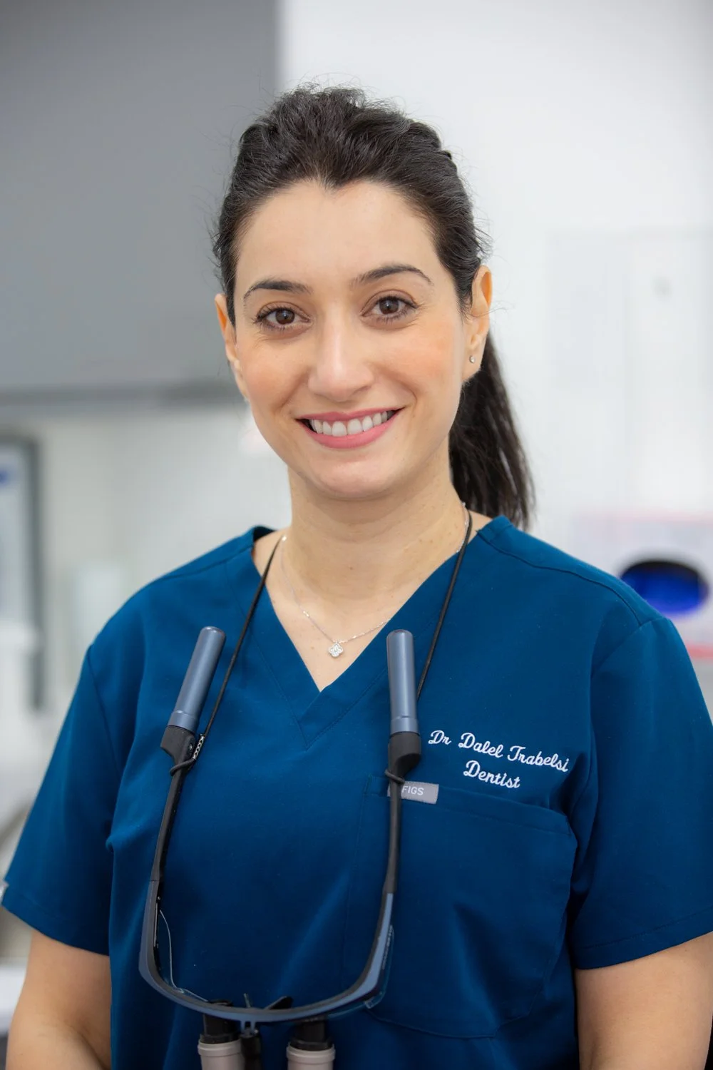 A smiling female dentist wearing blue scrubs and a dental headlamp, standing in a dental office.