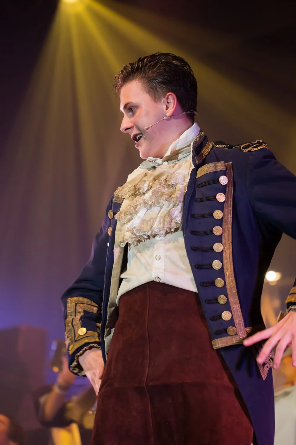A man dressed in a historical costume, resembling late 18th or early 19th-century military or aristocratic attire, performing on stage with a microphone headset, under stage lighting.