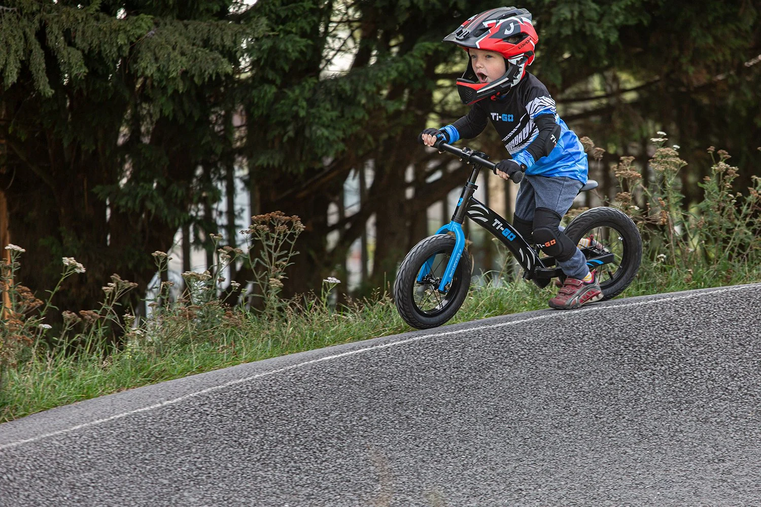 A young boy wearing a red helmet, black and blue jacket, gray pants, and riding shoes, riding a black and blue balance bike up a slight hill on a paved pathway with grassy area and dense trees in the background.