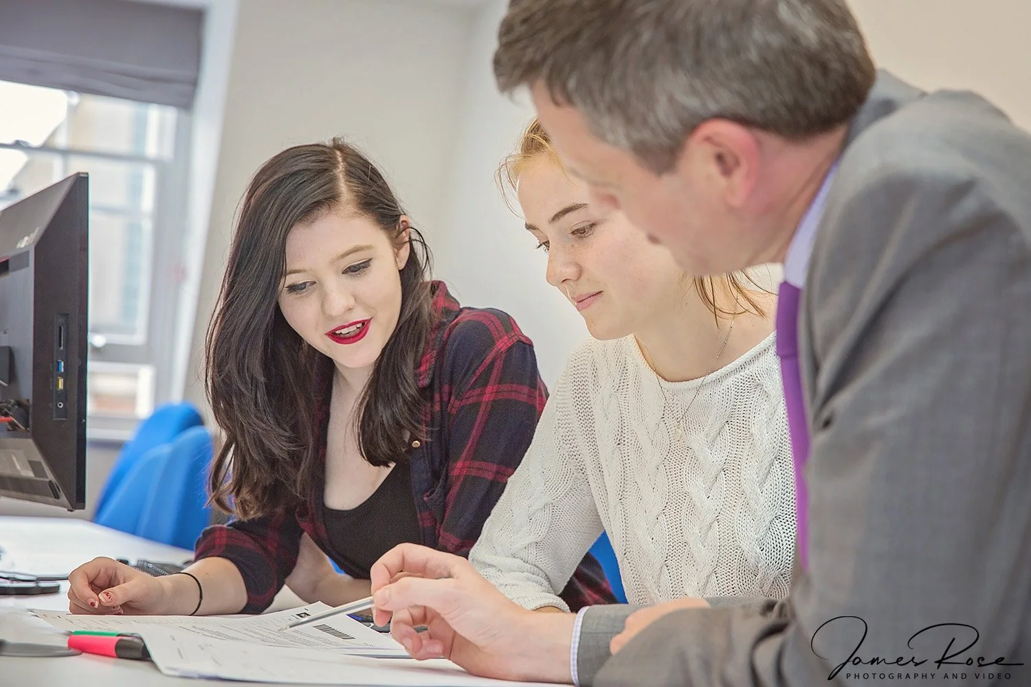 Three people in a business setting engaged in a discussion around documents on a table, with a computer monitor in the background.