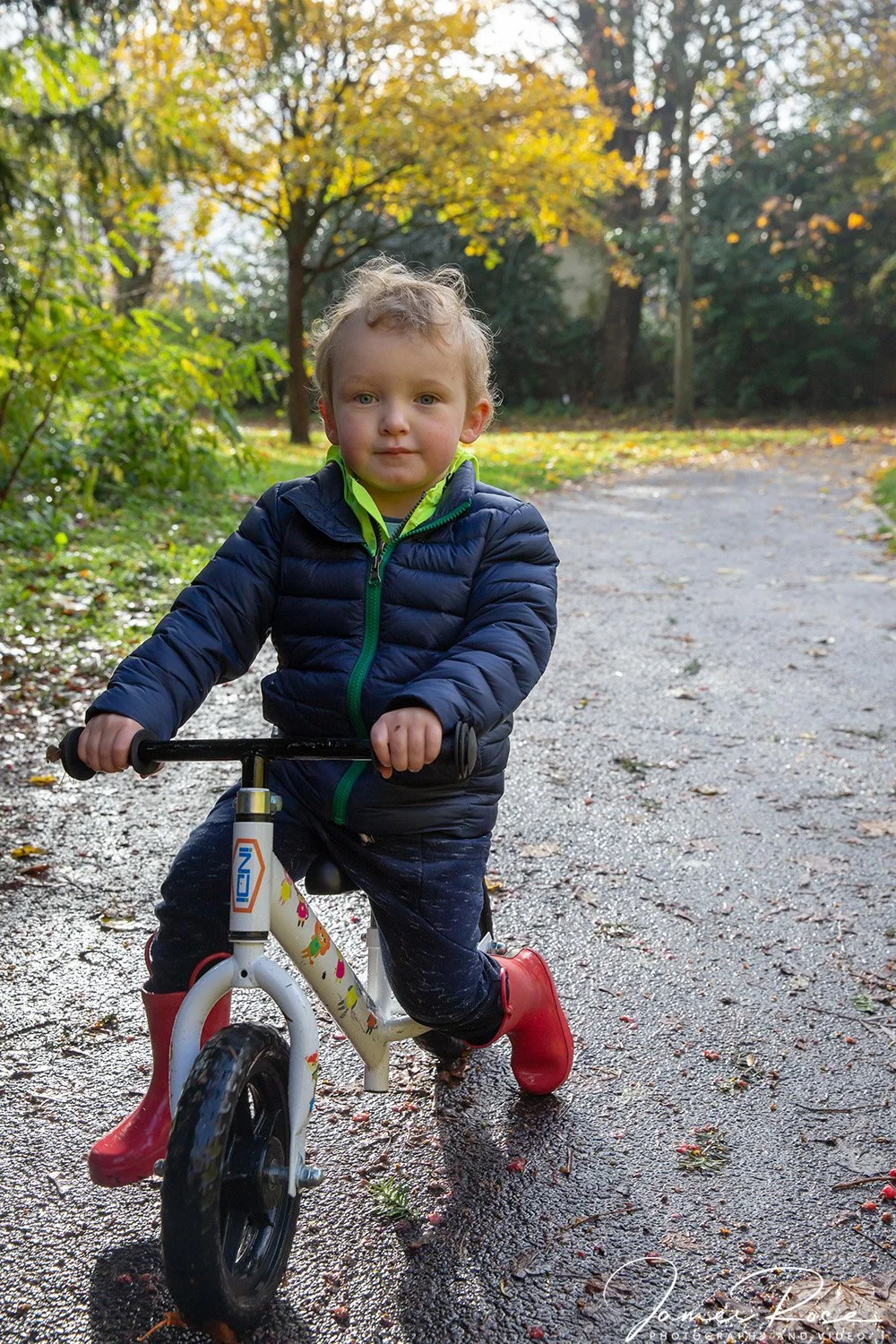 A young boy wearing a navy puffer jacket, dark pants with a pattern, and red shoes riding a small balance bike on a muddy, leaf-strewn pathway in a park during autumn, with trees exhibiting colorful fall foliage in the background.