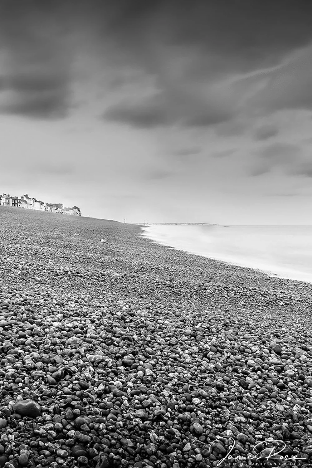 Black-and-white photo of a pebble beach with buildings in the distance, cloudy sky, and waves lapping the shore.