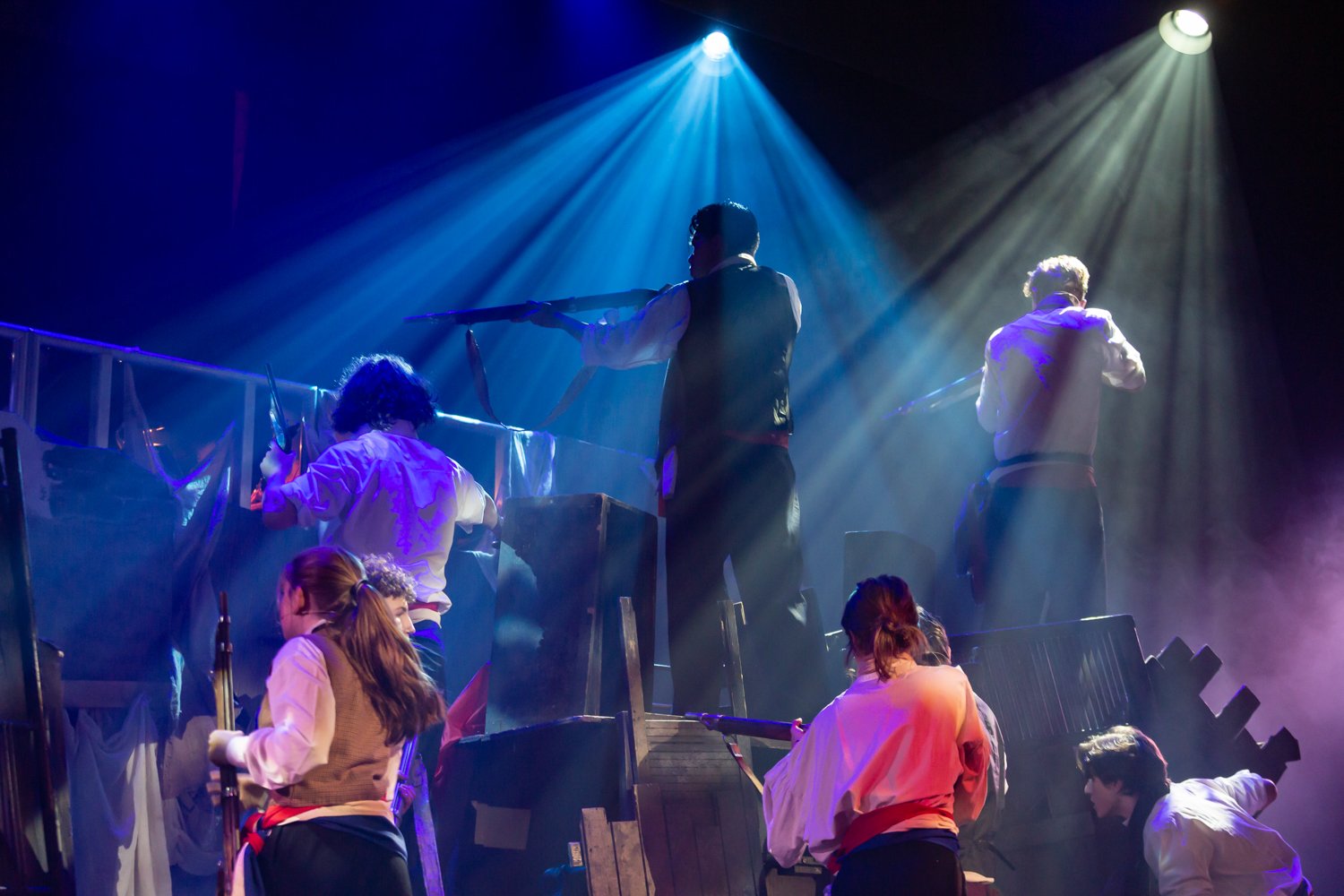 People performing a theatrical scene on stage with dramatic lighting and fog effects.