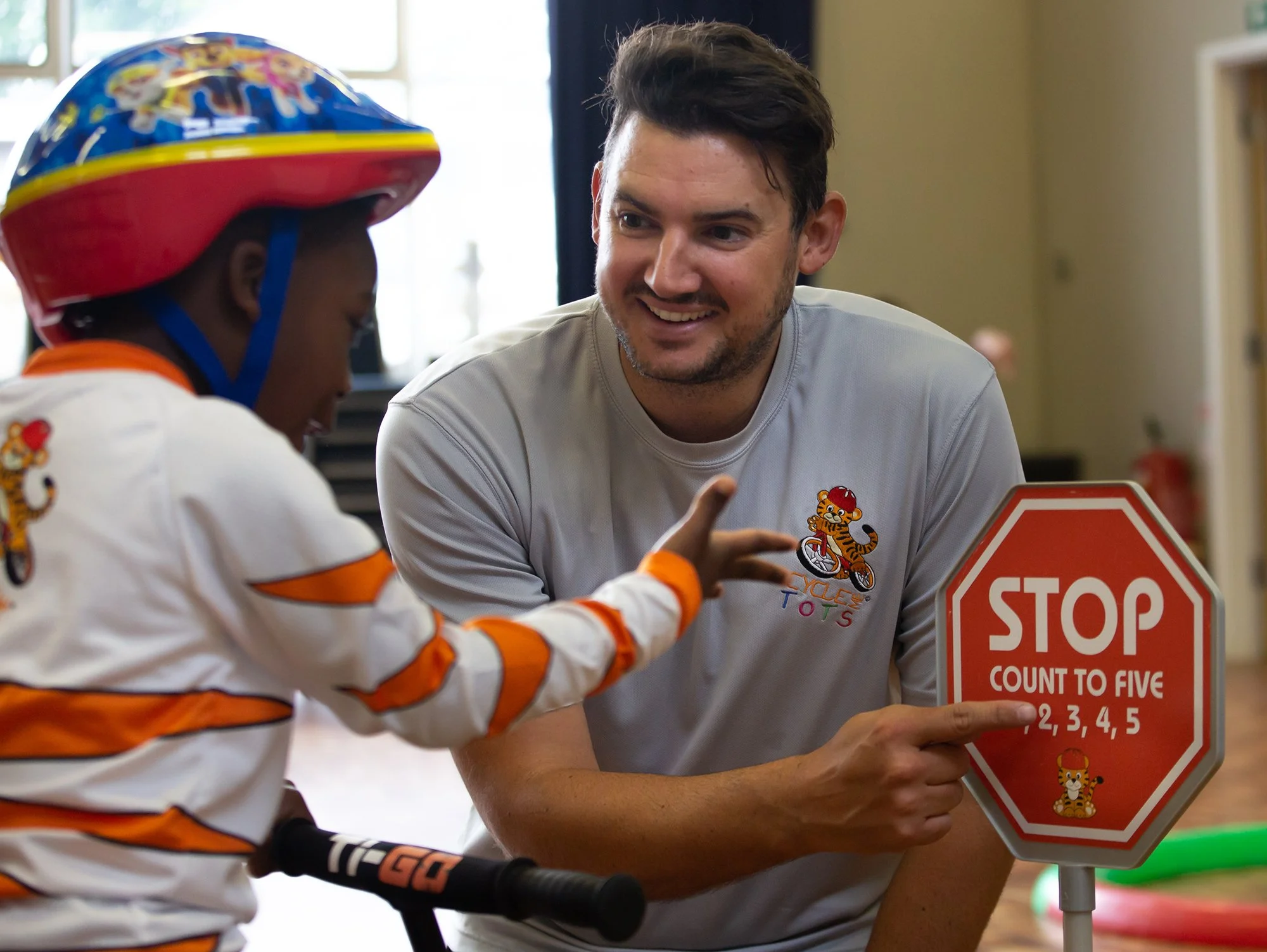 A smiling man in a gray T-shirt with a tiger and bicycle logo teaches a young boy, also wearing a similar T-shirt and a motorcycle helmet, to count to five using a stop sign.