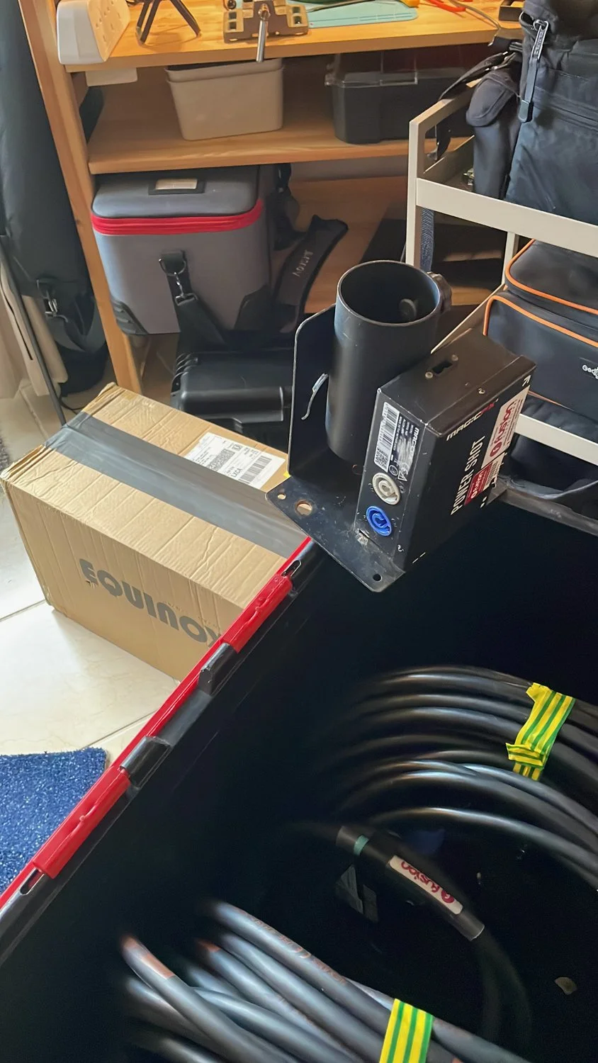 A cluttered garage workspace with various tools, equipment, and storage boxes, including a cardboard box, a black toolbox, a metal mount, and coiled cables.