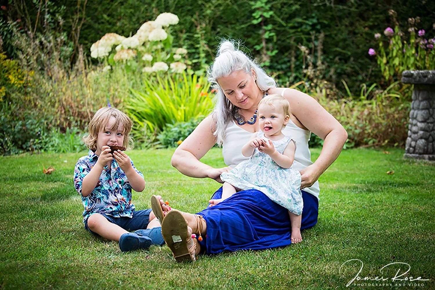 A woman with gray hair and children sitting on the grass in a garden, with one child eating a cupcake and another toddler sitting on her lap.