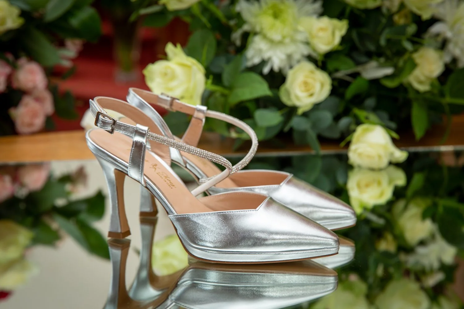 Silver high heel shoes with ankle straps on a reflective surface, with white and green flowers in the background.