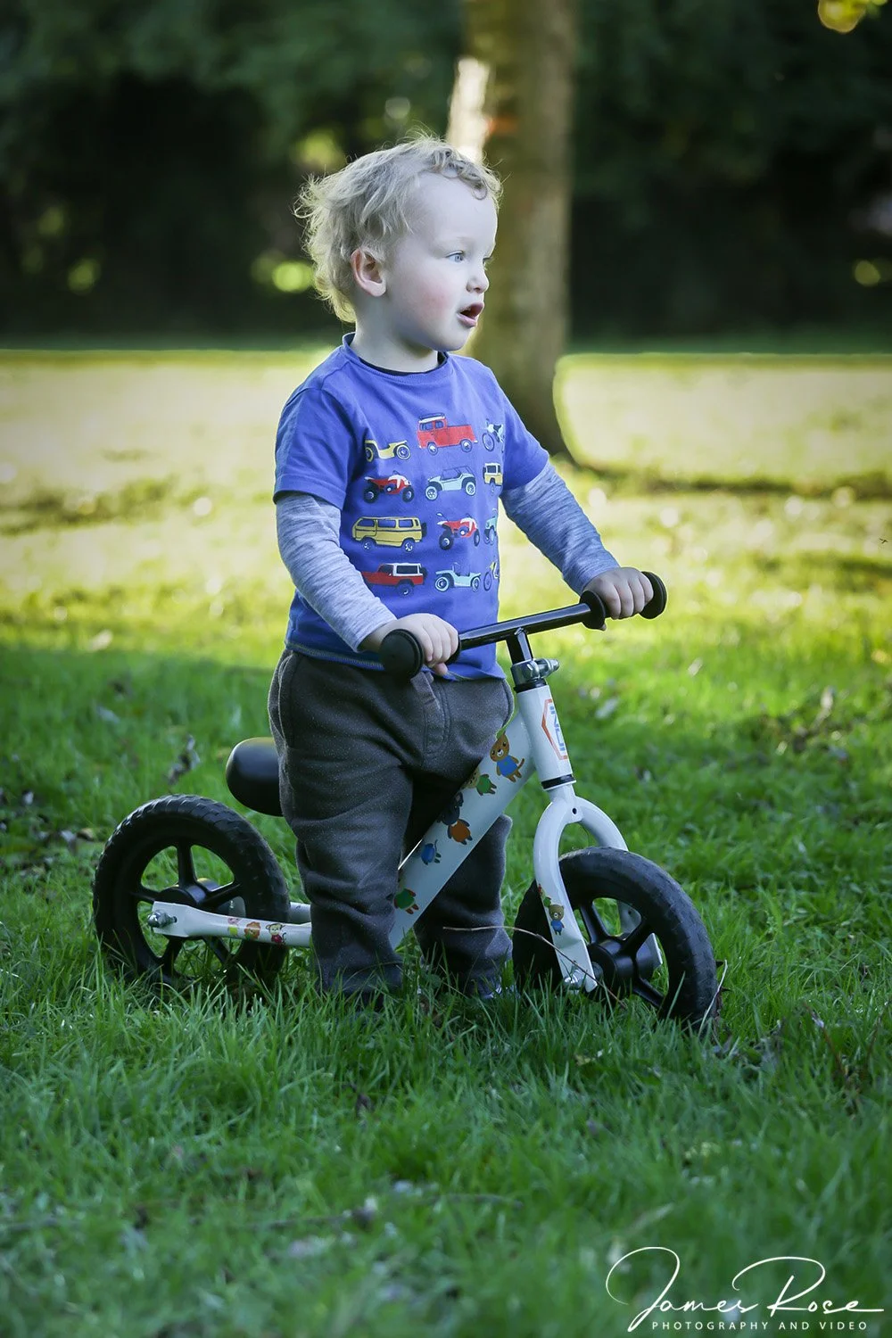 A young child with curly blonde hair riding a white balance bike outdoors on a grassy area, wearing a blue t-shirt with colorful vehicles and gray long sleeves underneath.